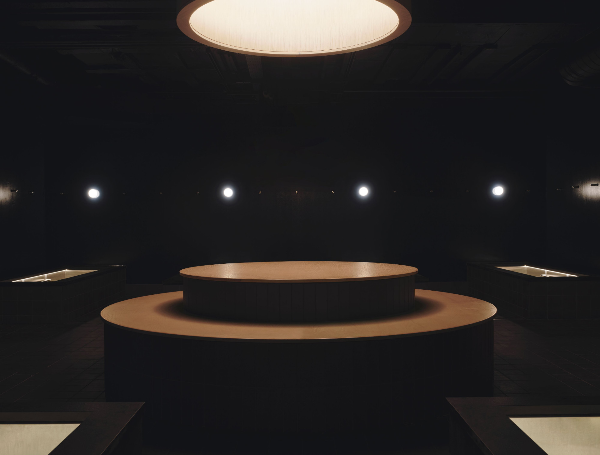 The dark, sparsely lit central lounge at ARC Canary Wharf, with the monolithic timber altar at its heart beneath a circular ceiling lightbox and four illuminated wall sconces