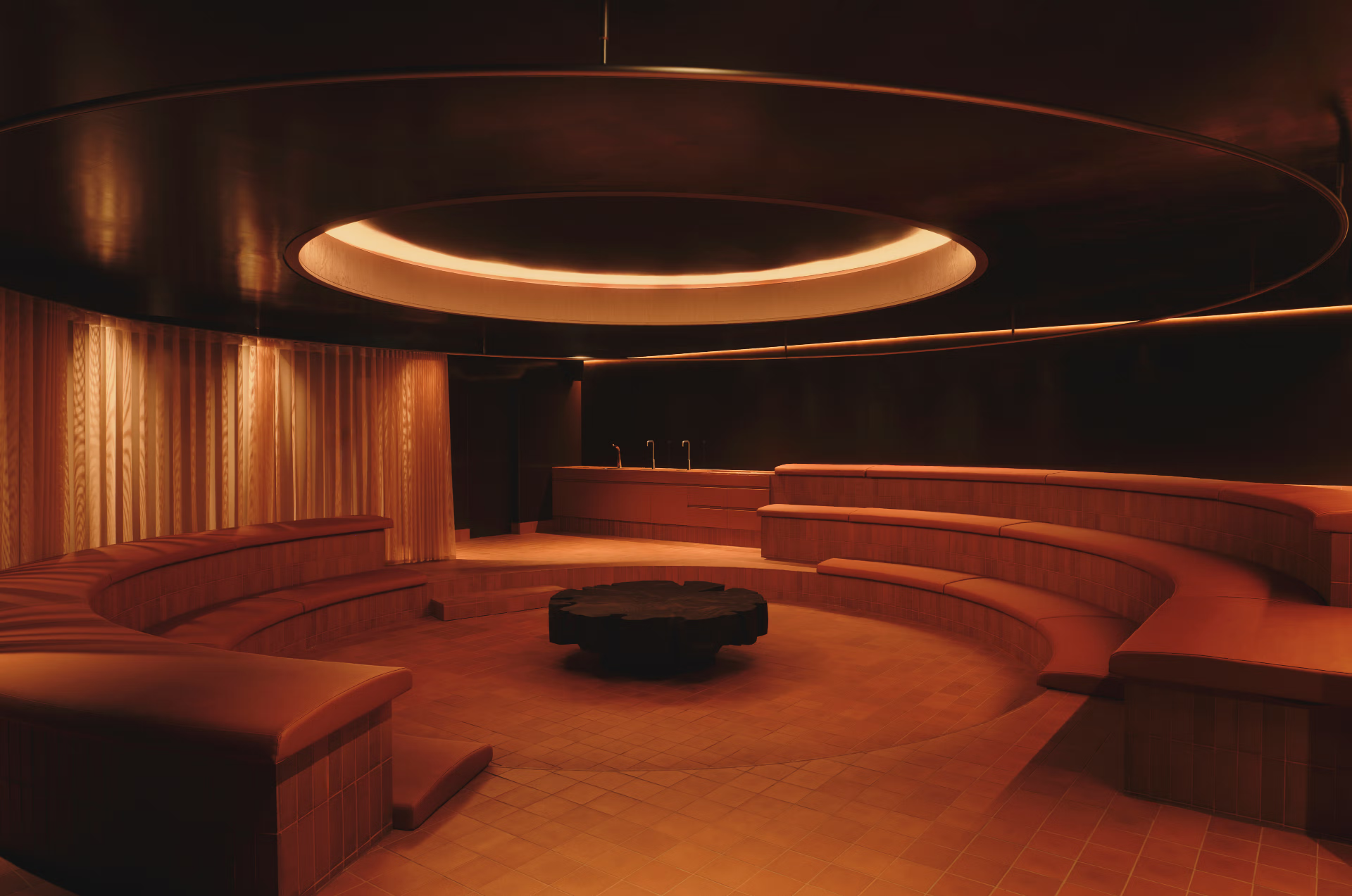ARC Canary Wharf's amphitheatre-style lounge — curved tiered terracotta-toned benches arranged around a central pit with a dark sculptural coffee table, beneath an oversized circular ceiling fixture
