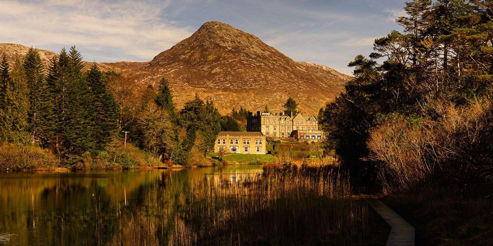 Ballynahinch Castle Hotel viewed across the Owenmore River at golden hour, with Benlettery and the 12 Bens mountain range rising behind it in Connemara, County Galway