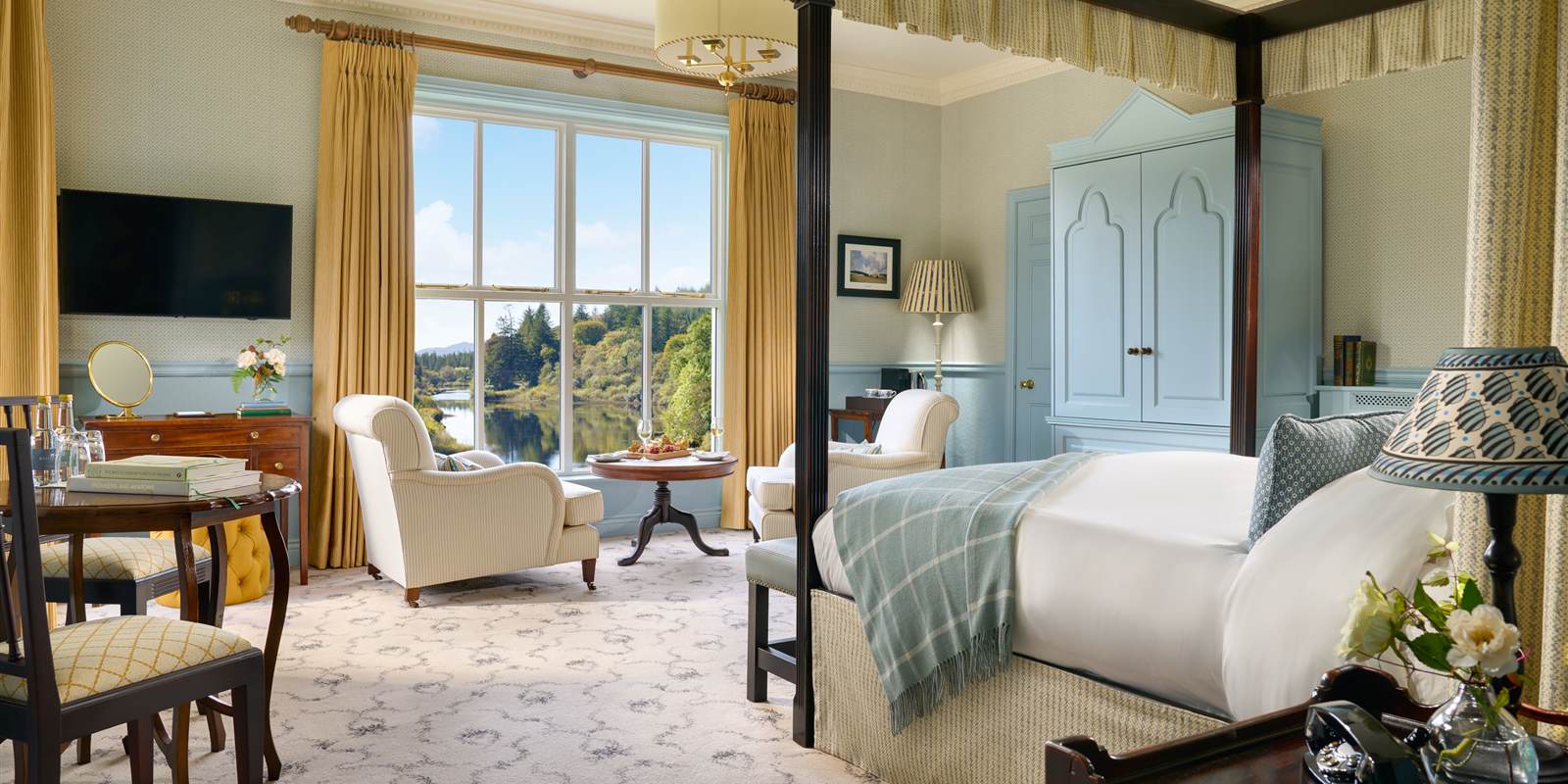 Superior bedroom at Ballynahinch Castle Hotel — four-poster bed, hand-painted wardrobe and tall sash window framing the Owenmore River and Connemara woodland
