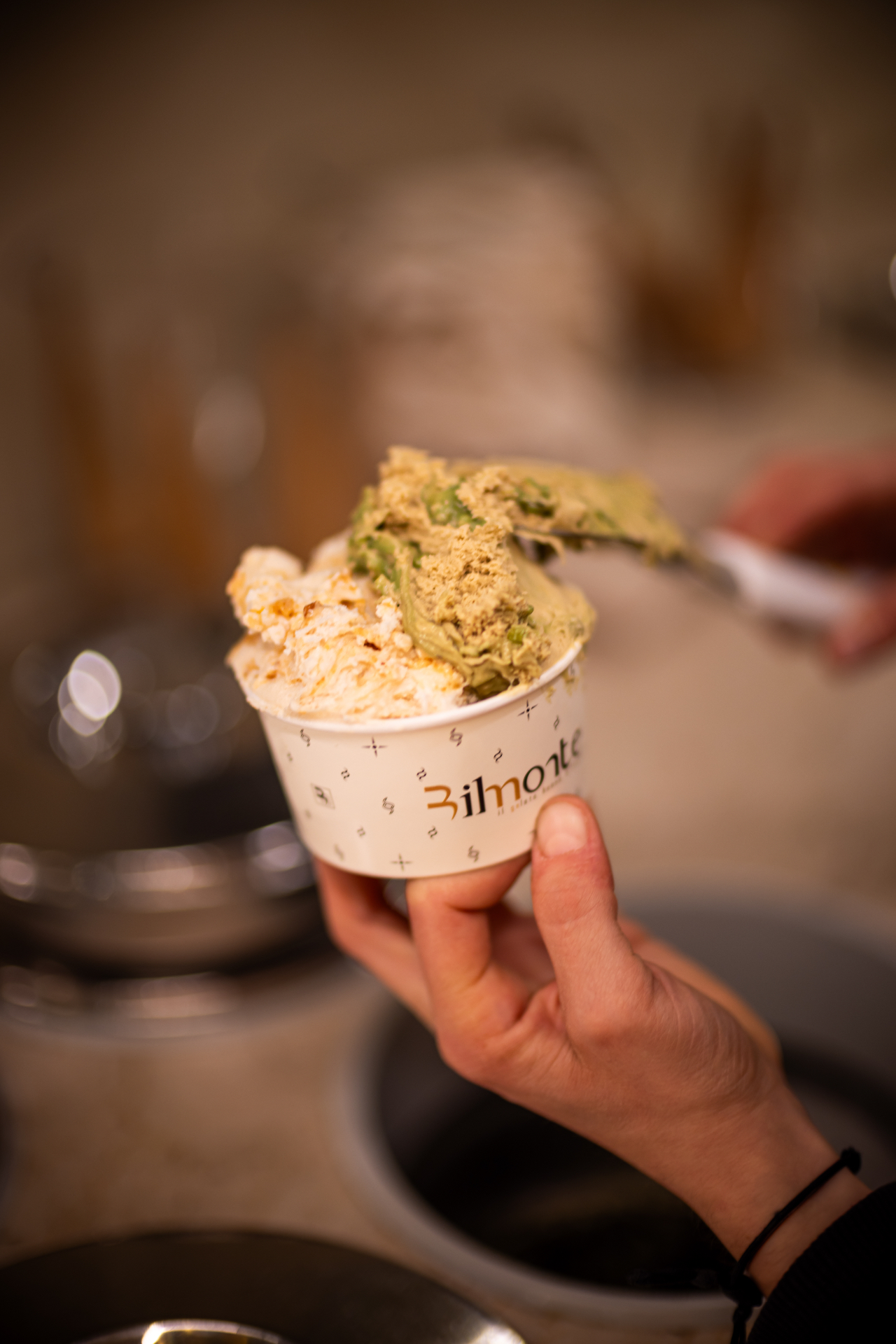 Hand holding a Bilmonte cup of pistachio and hazelnut gelato with a spoon