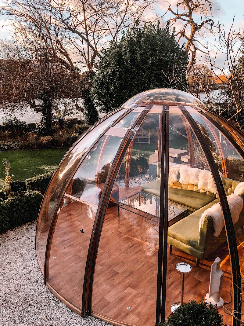 A copper-framed glass dining dome in the garden at Bingham Riverhouse, Richmond, at dusk, with sheepskin-throw sofas inside and the Thames visible through bare winter trees behind