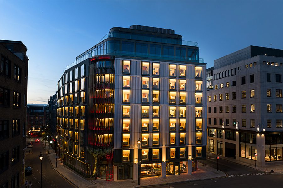 The BoTree hotel, Marylebone — the illuminated exterior at dusk, showing the corner building's warm-lit windows and a red vertical latticed detail on the facade that echoes the diamond pattern of the demolished Welbeck Street car park