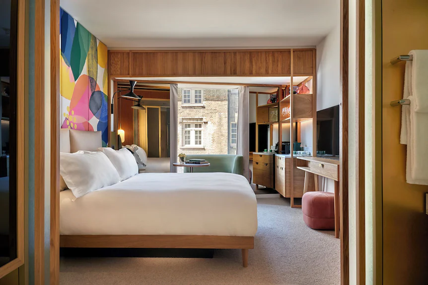 A suite at The BoTree — a linen-dressed bed against a colourful abstract mural, warm wood joinery, a green armchair by the window and a dressing table with a soft pink stool
