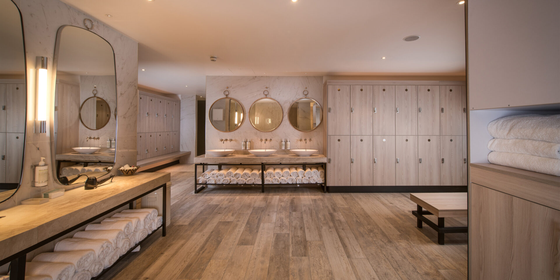 BXR Marylebone — the members' changing room with marble walls, round brass-rimmed mirrors, timber lockers and rolled white towels stacked on stone shelves