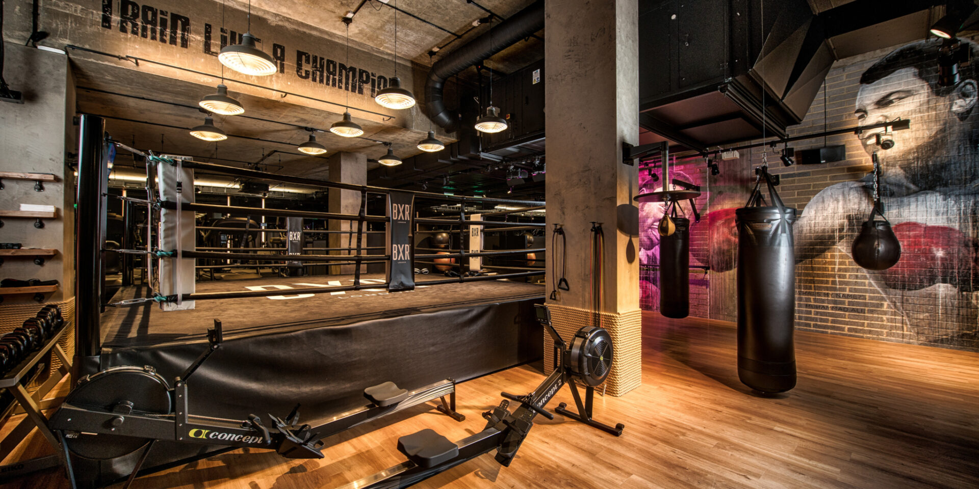 BXR Marylebone — the full-size boxing ring beneath 'Train Like a Champion' stencilling, warm pendant lighting, Concept2 rowers on timber flooring and a Muhammad Ali mural on brick