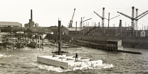 A black-and-white photograph from the 1940s showing the original Bindon Blood Stoney diving bell in operation in Dublin Port, a workman standing on a precast concrete block beside the bell in the River Liffey