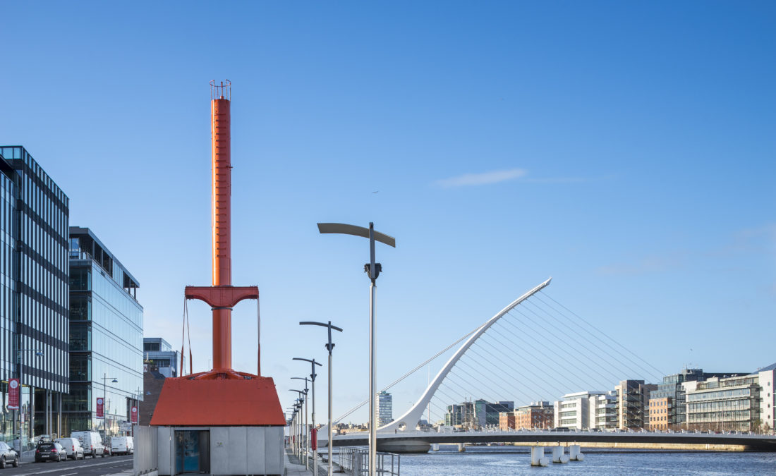 The Diving Bell standing on Sir John Rogerson's Quay in daylight, the Samuel Beckett Bridge spanning the Liffey behind it, modern Docklands office buildings on either side