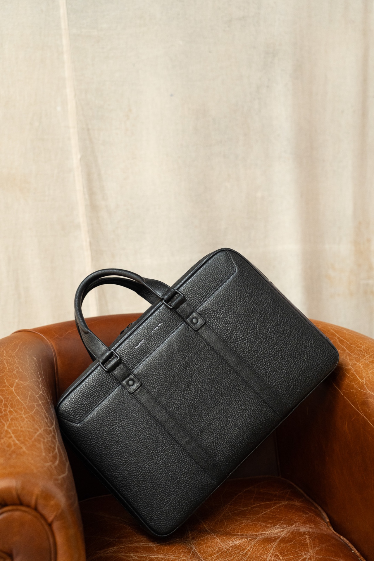 Grams28 black full-grain leather briefcase resting on a brown leather armchair