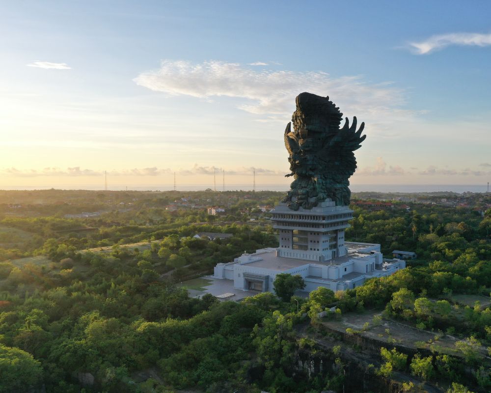 Garuda Wisnu Kencana statue at dawn — aerial view above the limestone hills of the Bukit Peninsula with the Indian Ocean beyond