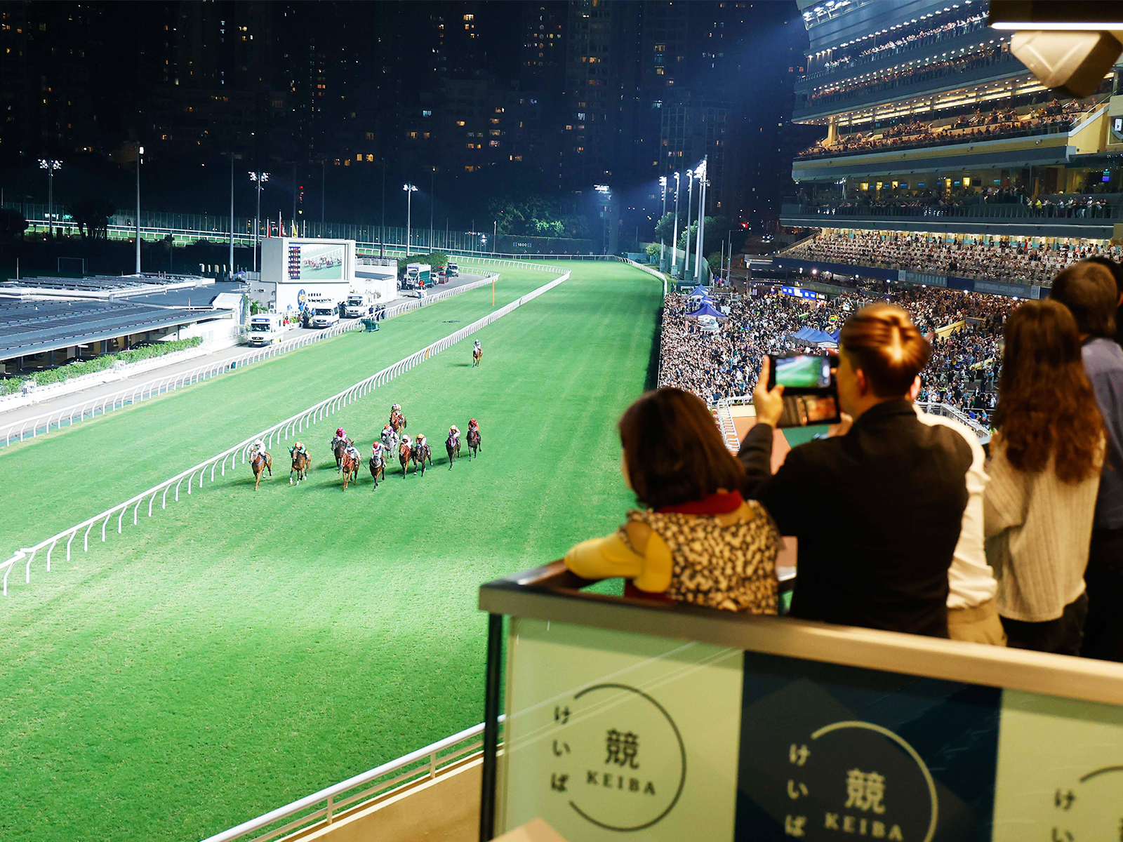 Spectators on a high terrace photographing the field thundering up the floodlit Happy Valley finishing straight at night, with the grandstand and city skyline glowing on the right