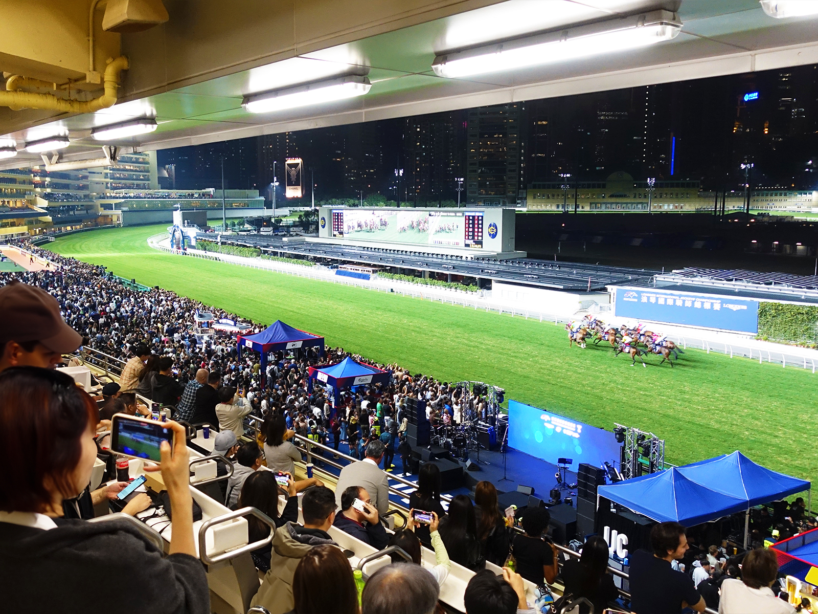 Race night at Happy Valley seen from a 2F terrace — packed grandstand on the left, horses thundering down the home straight on the right, illuminated Wan Chai towers behind