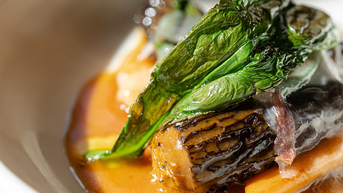 A close-up plated dish at Lita — a caramelised, grill-marked portion of fish and vegetable set in a burnt-orange sauce, topped with a glossy charred green leaf and cured pork, glistening under warm light