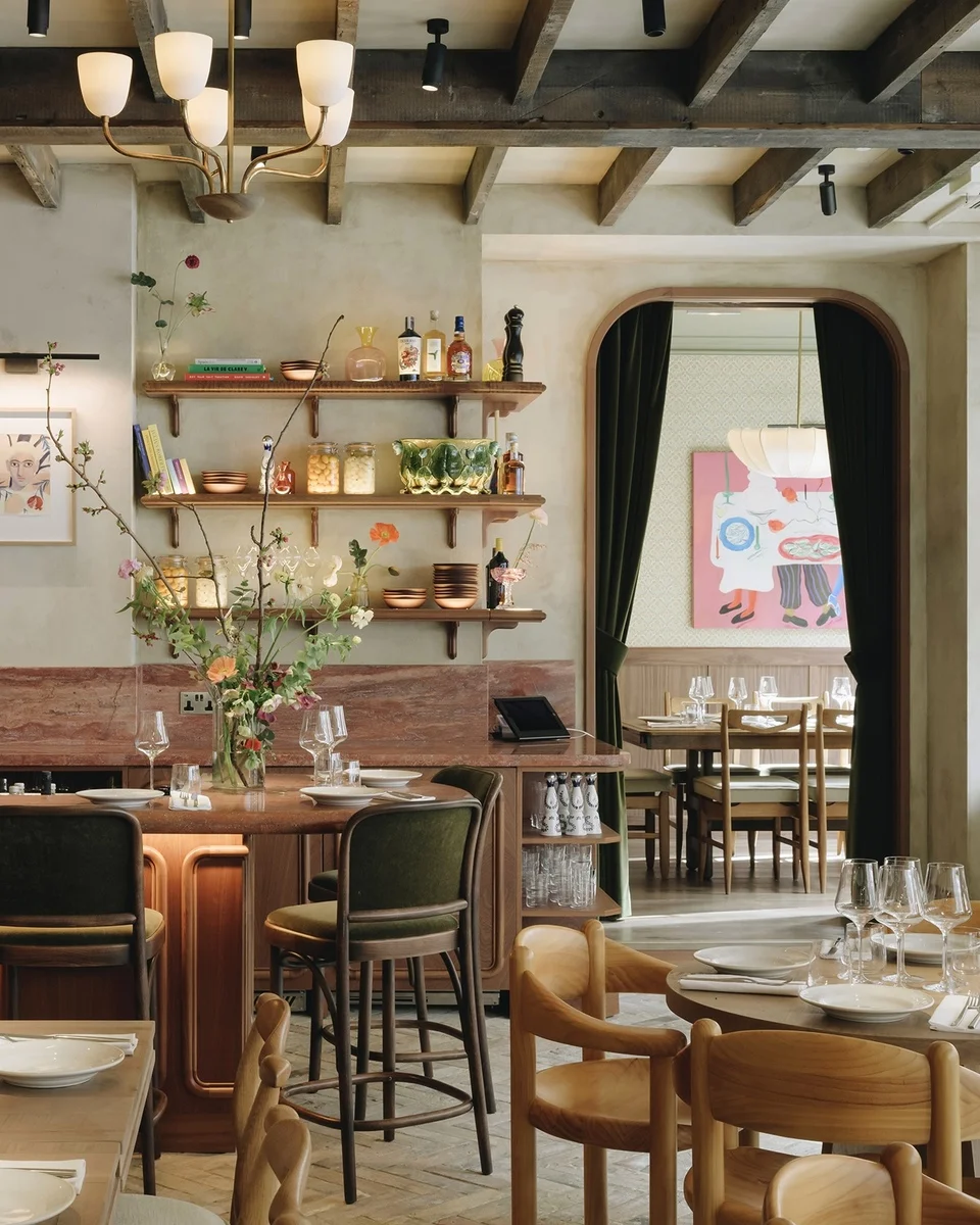 Lita's dining room — brass chandeliers under exposed wooden beams, open kitchen shelves lined with bottles and books above a rust-red marble bar, spring flowers in tall glass vases, green velvet bar stools and warm timber floors