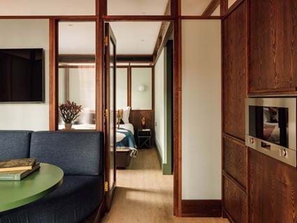 A Locke Canary Wharf one-bedroom suite — a curved navy sofa with a round green coffee table in the foreground, a wall-mounted television, and a view through to the bedroom and kitchen beyond the reeded-glass partitions