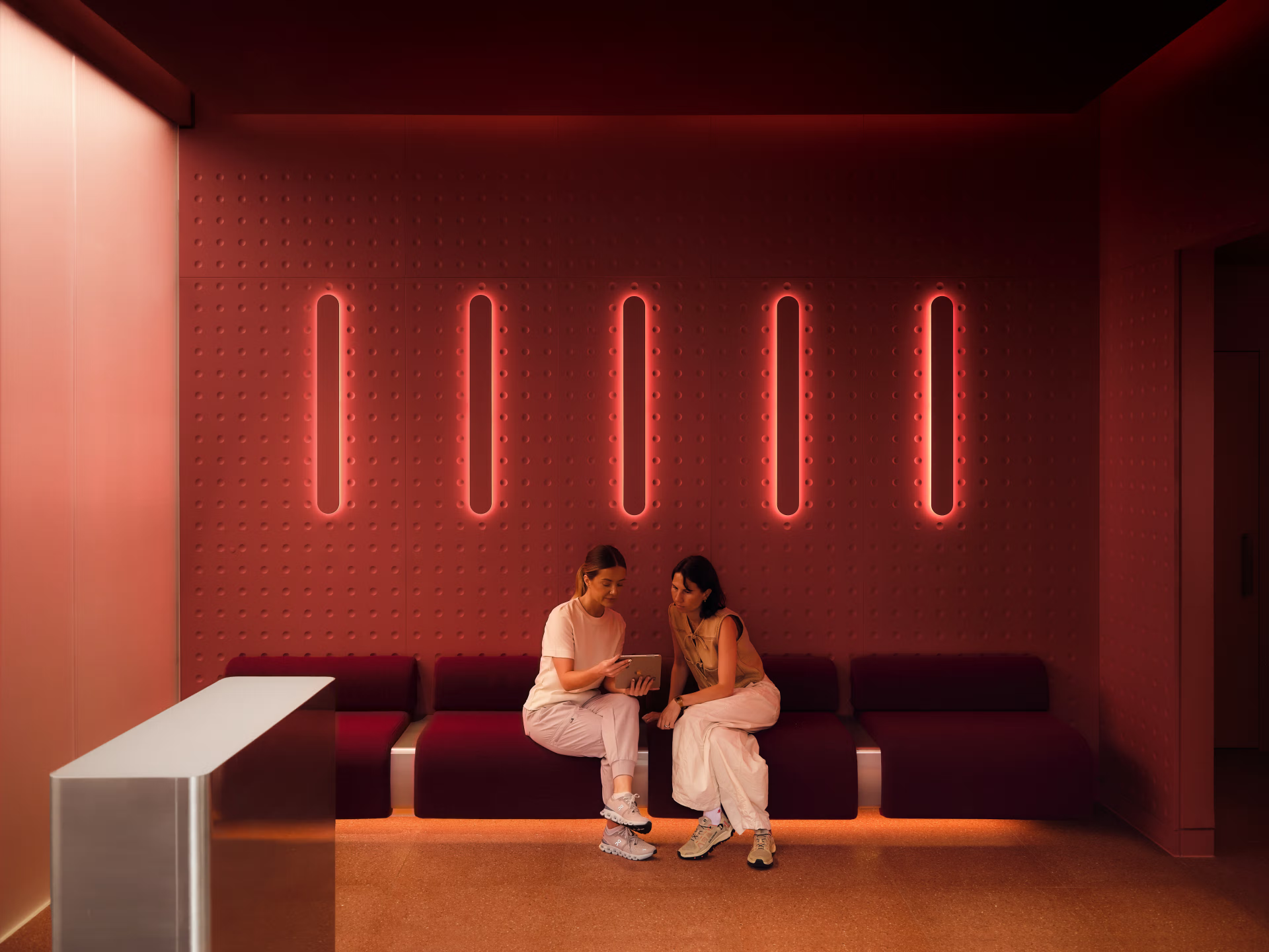 Neko Health clinic waiting lounge — deep red walls with vertical neon light panels and clients reviewing results on a tablet