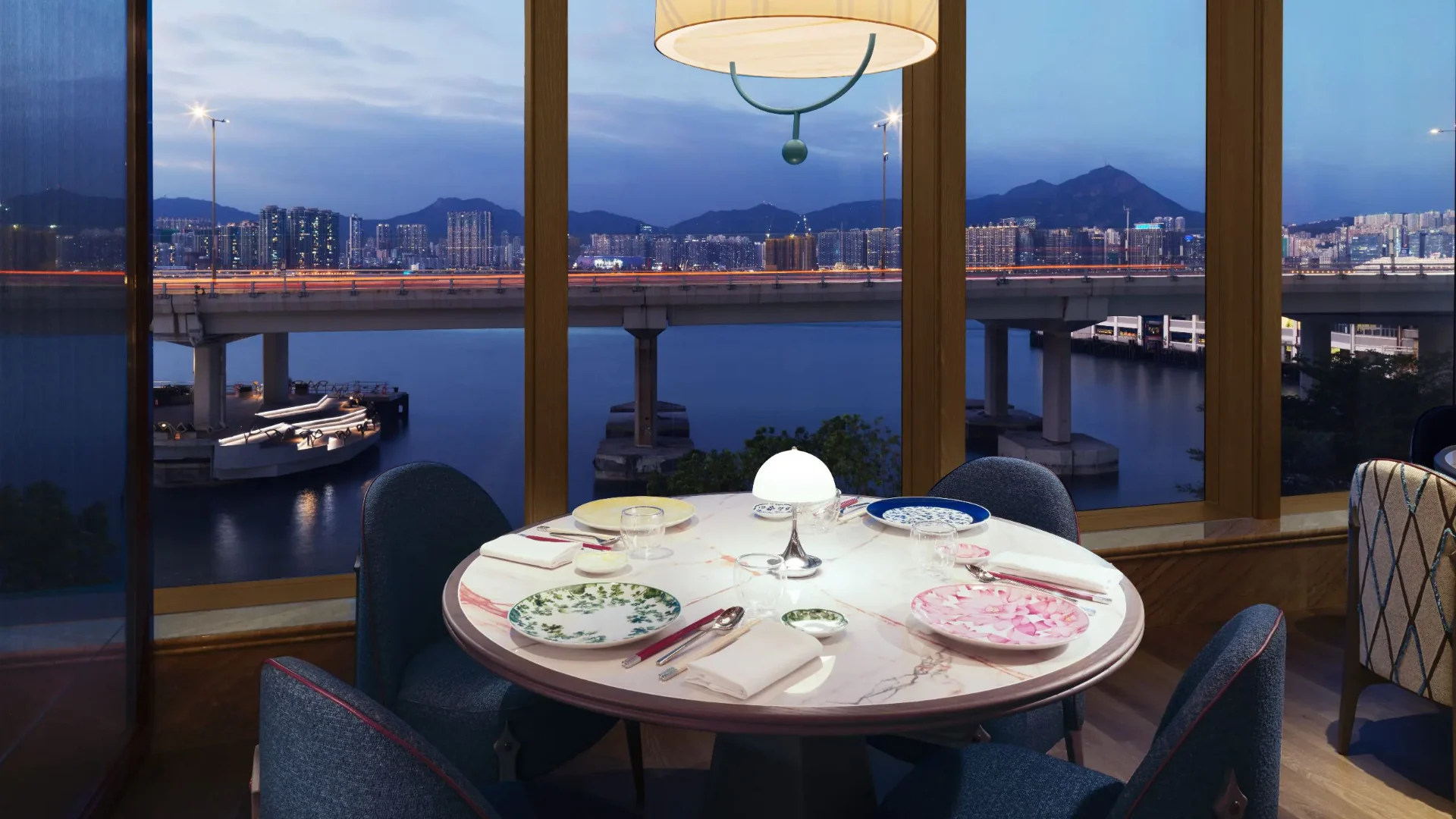 A round dining table set for four at One Duck Lane with the Victoria Harbour skyline and bridge lights visible through floor-to-ceiling windows at dusk