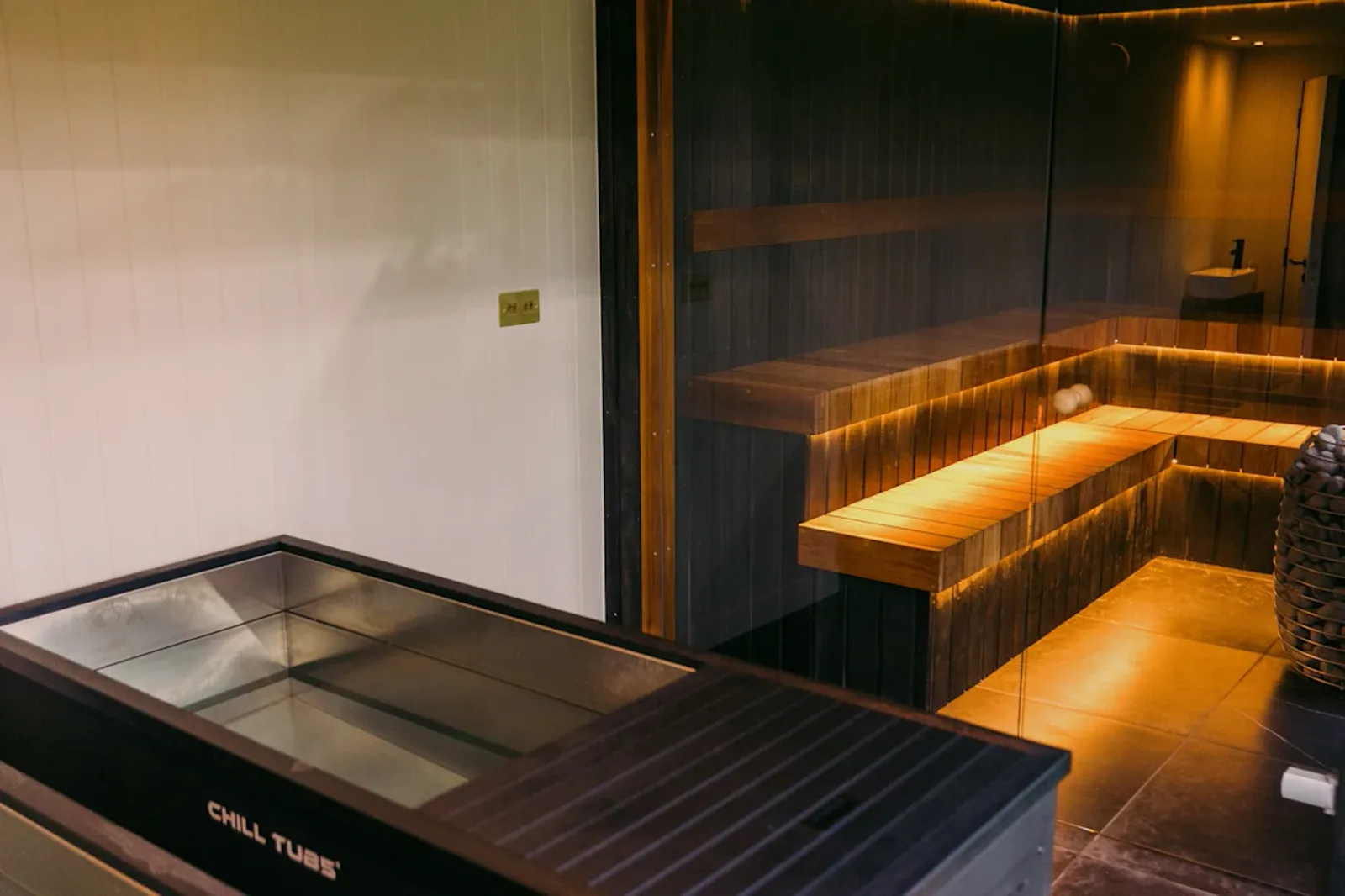 Padel Social Club Earl's Court — the wellness area, a warm-lit sauna cabin beside a stainless steel cold plunge