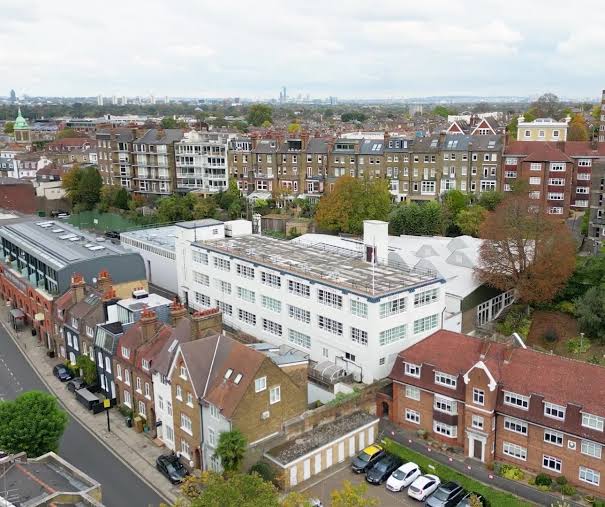 An aerial view of the Poppy Factory building in Richmond — a white, low-rise industrial building with a distinctive sawtooth roof, tucked among Victorian mansion blocks, brick houses and mature trees, with the London skyline in the distance