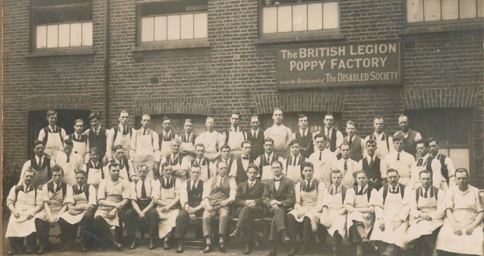 A sepia photograph of Poppy Factory workers in aprons and ties gathered outside the original British Legion Poppy Factory building, under a sign reading 'The British Legion Poppy Factory under the Management of The Disabled Society'