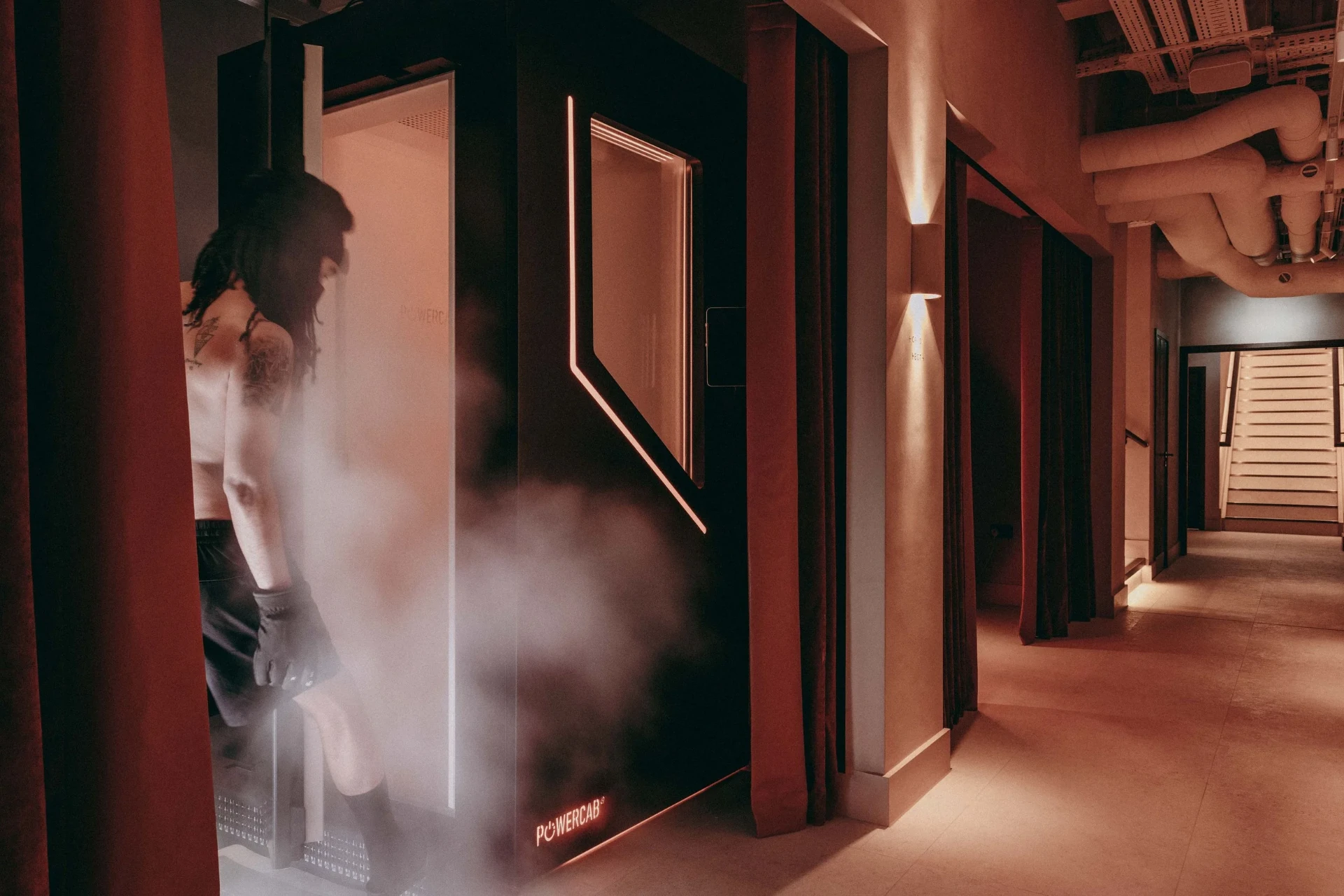 Rebase Recovery — the cryotherapy chamber viewed from a warm-lit corridor, with a member preparing to step inside