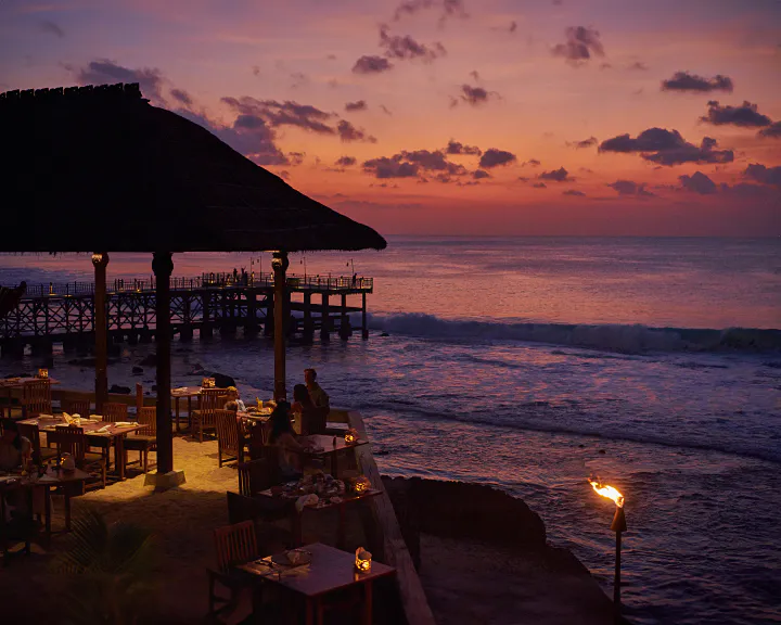 Rock Bar at AYANA at dusk — thatched-roof pavilion and tiki torches with a long pier silhouette against the pink Jimbaran sunset