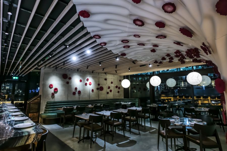 Roe's main dining room with a sculptural white draped ceiling installation scattered with deep red coral-like forms, concrete walls dotted with crimson floral pieces, dark green banquettes and pendant globe lights
