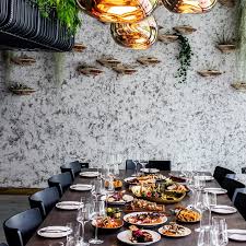 A long sharing table at Roe set for a group, scattered with plates of food and lined with dark chairs, beneath a stone-textured wall lined with bronze fish sculptures and warm amber pendant lights overhead