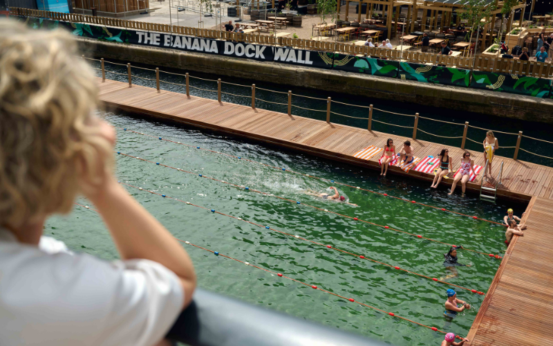 Deck view at Sea Lanes Canary Wharf — a woman watches swimmers in the lane pool while bathers sit along the boardwalk edge, with the dock-side restaurant terraces beyond