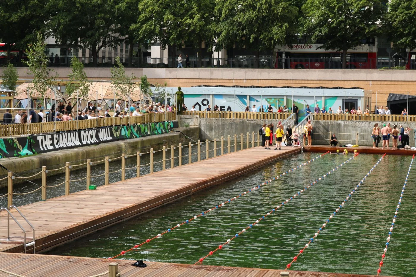 Sea Lanes Canary Wharf on opening day — wide view of the six-lane floating pool with swimmers, the timber boardwalk and 'The Banana Dock Wall' graphic across the quayside, geodesic domes and a London bus visible above