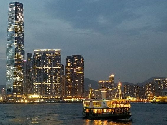 A Star Ferry illuminated at dusk on Victoria Harbour, with the lit Tsim Sha Tsui skyline and the International Commerce Centre tower behind it