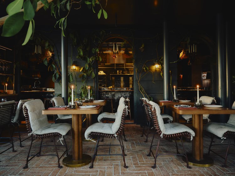Tower House, Richmond — the candlelit main dining room with dark walls, iron pillars trailing with ivy and vines, brick floor, and wooden tables set with linen cushions and taper candles