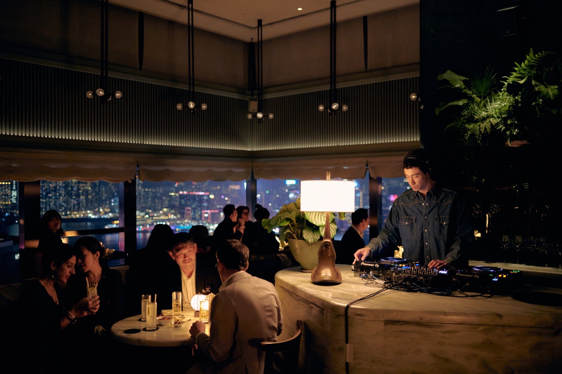 The Green Room bar at The Upper House at night with a DJ at the marble counter, low lamps and the Kowloon skyline glowing through the windows behind