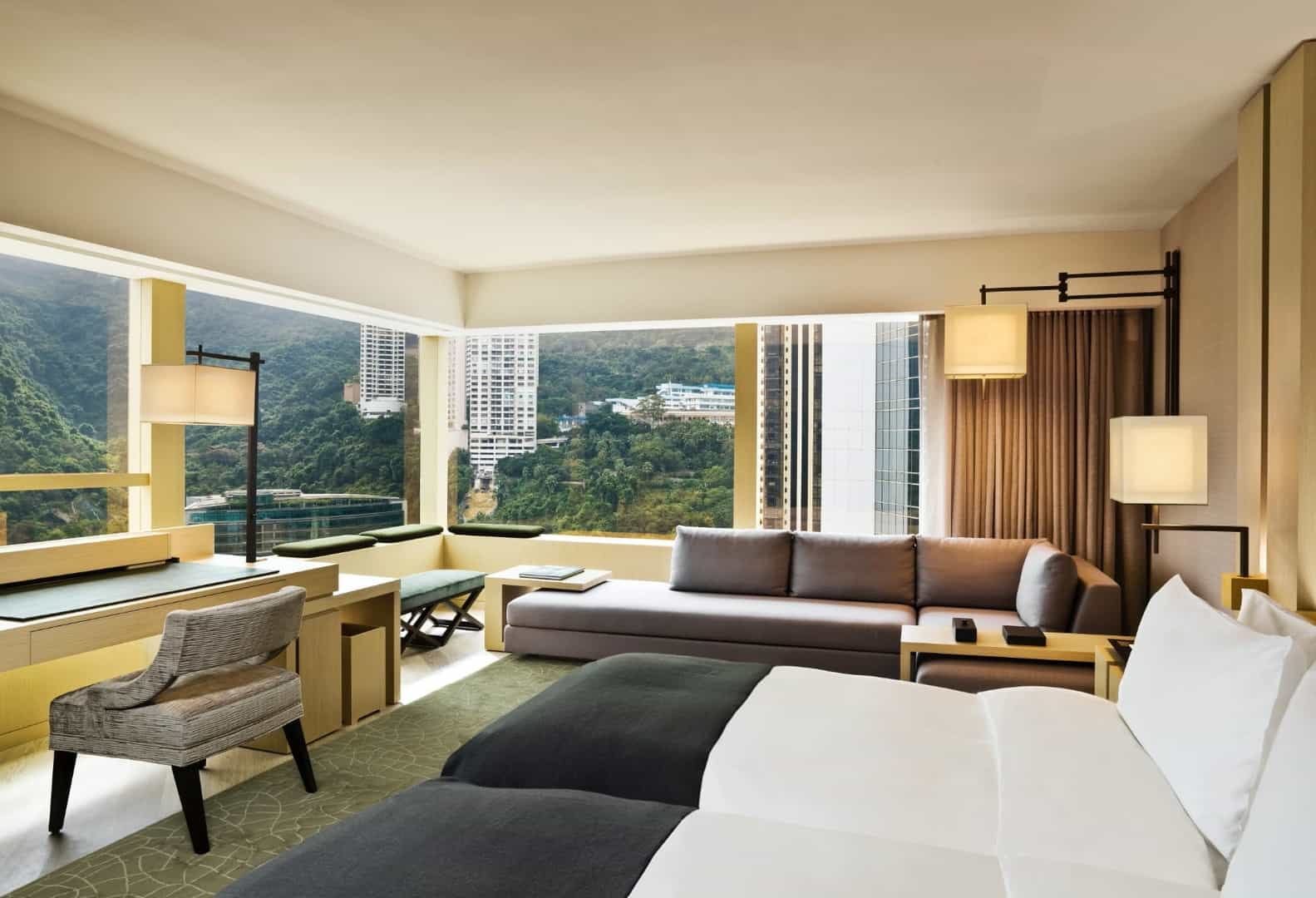 An Upper House room with corner windows looking onto Hong Kong Island's green hillsides and adjacent towers, oak and bamboo finishes, low sofa and writing desk