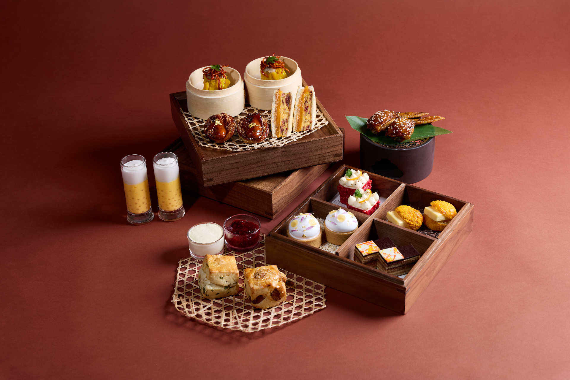 An afternoon tea spread at Salisterra served in wooden bamboo-trayed tiers against a deep red backdrop — dim sum, scones, glazed wings, mango pudding shots and patisserie