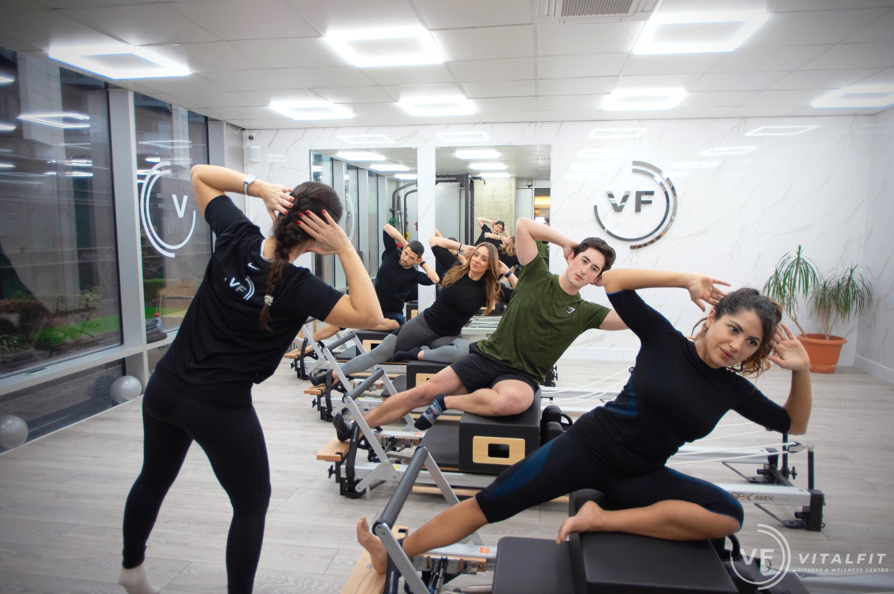 A reformer Pilates class in session at VitalFit Canary Wharf — clients on reformer beds following an instructor through a side-bend sequence, set against the studio's marble feature wall with the VF logo