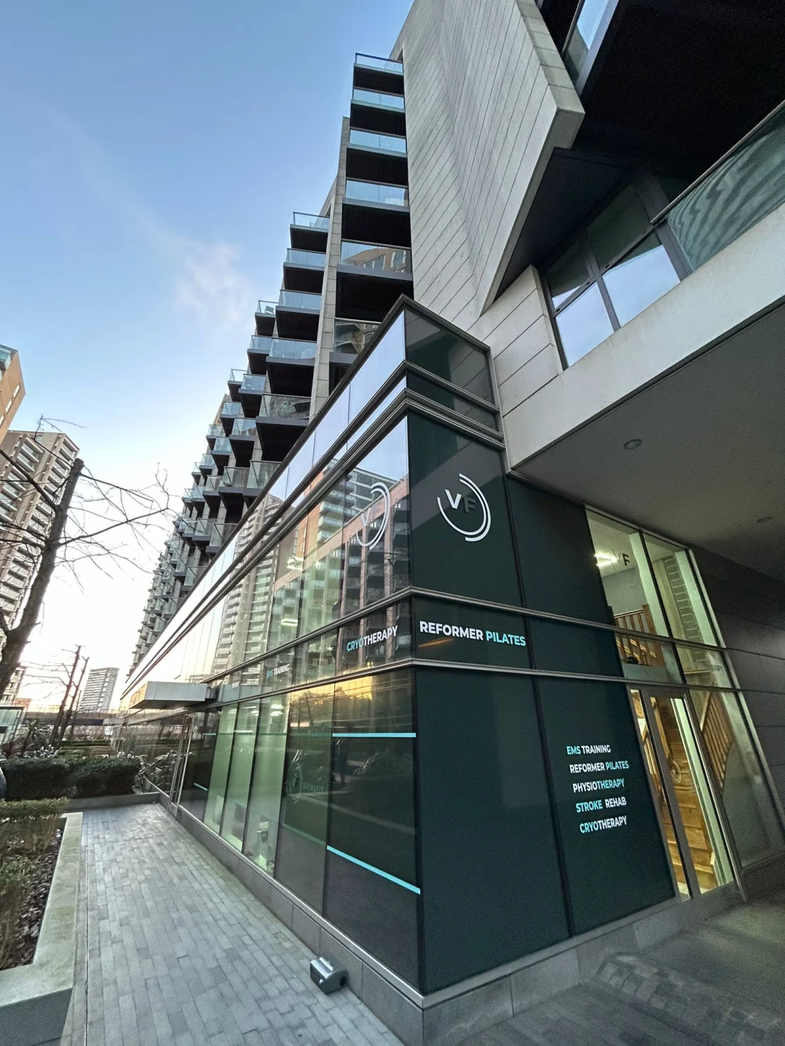 The exterior of VitalFit at Baltimore Wharf, Canary Wharf — a glass-fronted ground-floor unit with a deep green panel bearing the VF logo, with service signage reading EMS Training, Reformer Pilates, Physiotherapy, Stroke Rehab and Cryotherapy, set beneath a residential tower