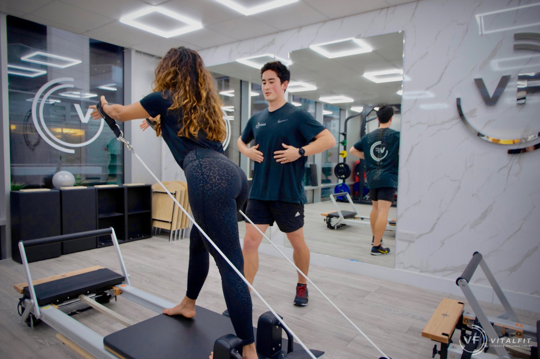 A VitalFit instructor in a dark VF t-shirt coaching a client through a standing reformer exercise, in a small, mirrored studio with a marble feature wall and the VF circular logo etched into the glass partition
