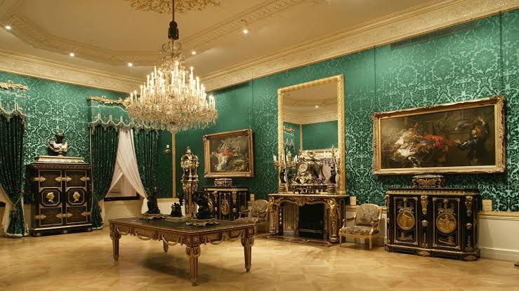 A green damask-walled room at the Wallace Collection with a crystal chandelier, gilded French furniture and Old Master paintings