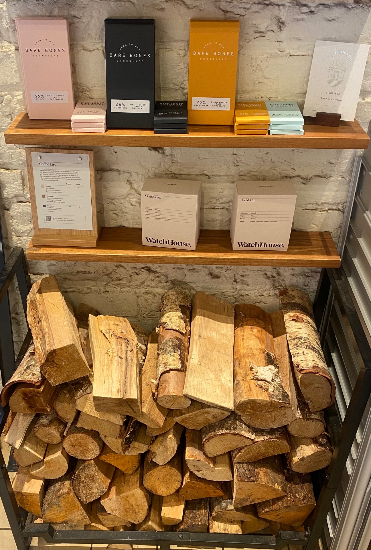 WatchHouse retail shelves with Bare Bones chocolate, coffee bags, and firewood against white brick wall