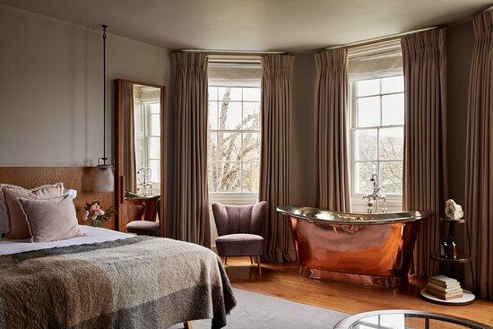 A bedroom at Bingham Riverhouse, Richmond — muted taupe curtains at tall Georgian sash windows, a copper roll-top bath beside the bed, and pale linens with a soft grey throw