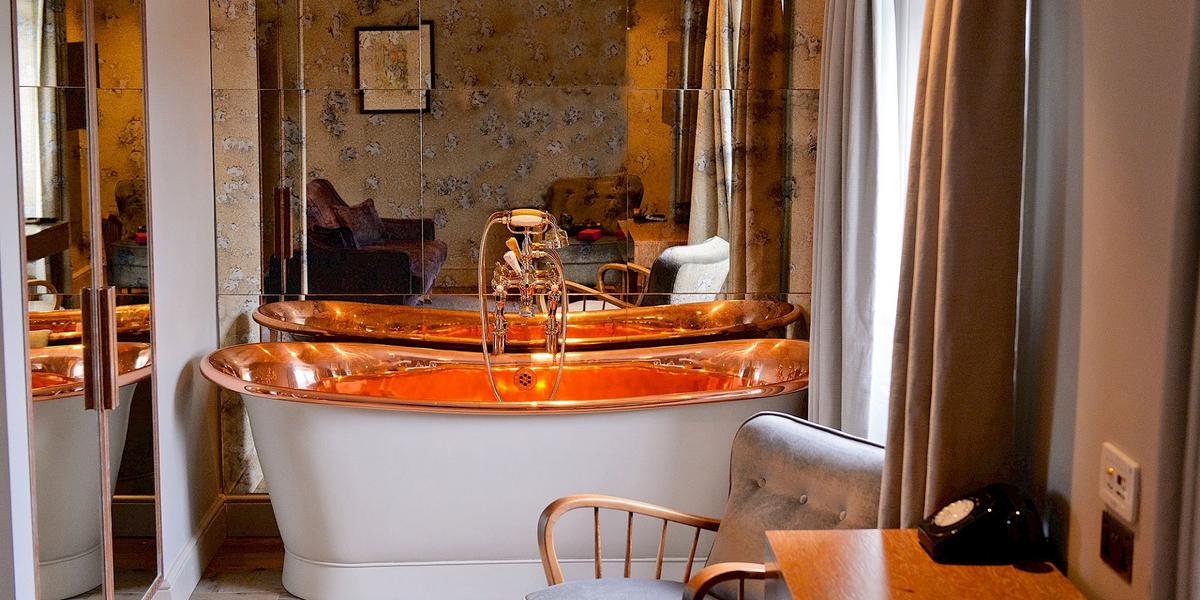 A polished copper roll-top bath at Bingham Riverhouse against an antiqued mirror wall and painterly wallpaper, with a small vintage phone on a side table