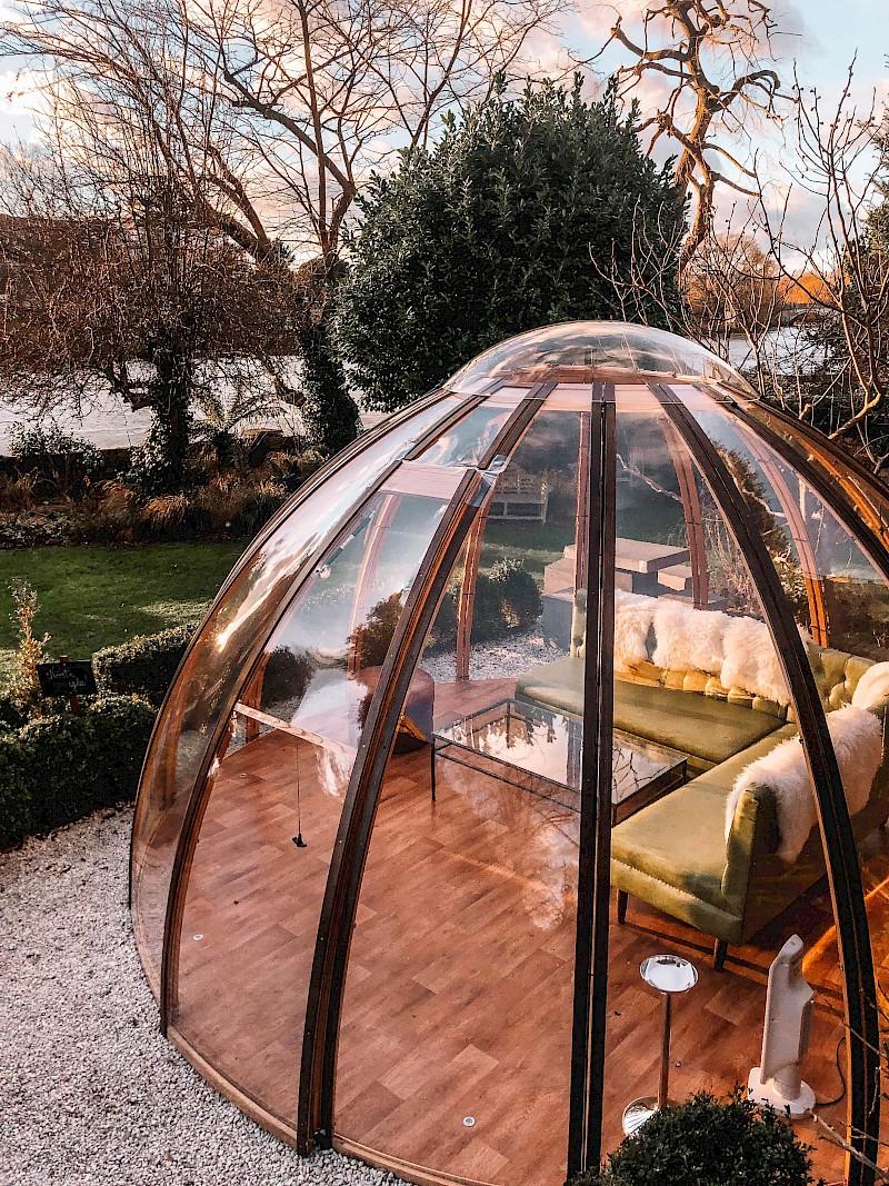 A copper-framed glass dining dome in the garden at Bingham Riverhouse, Richmond, at dusk, with sheepskin-throw sofas inside and the Thames visible through bare winter trees behind