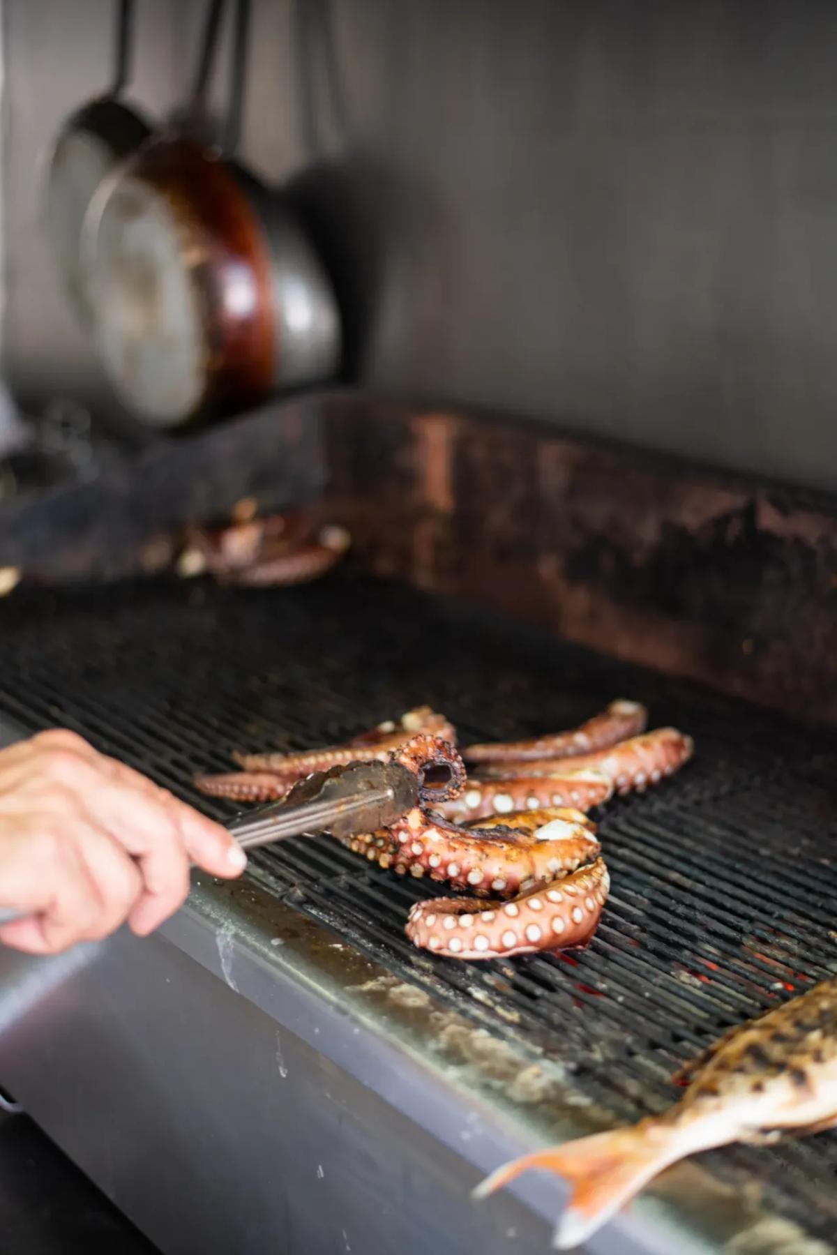 Octopus and whole fish grilling over charcoal at Drakos Taverna, tongs turning tentacles on the grill