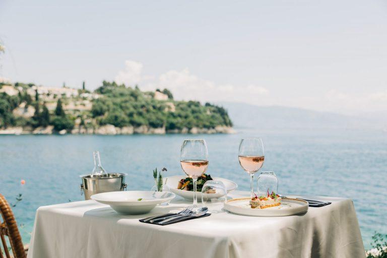 Waterside table at The White House, Kalami — white linen, rosé wine and plated food set against the open sea and Albanian coastline