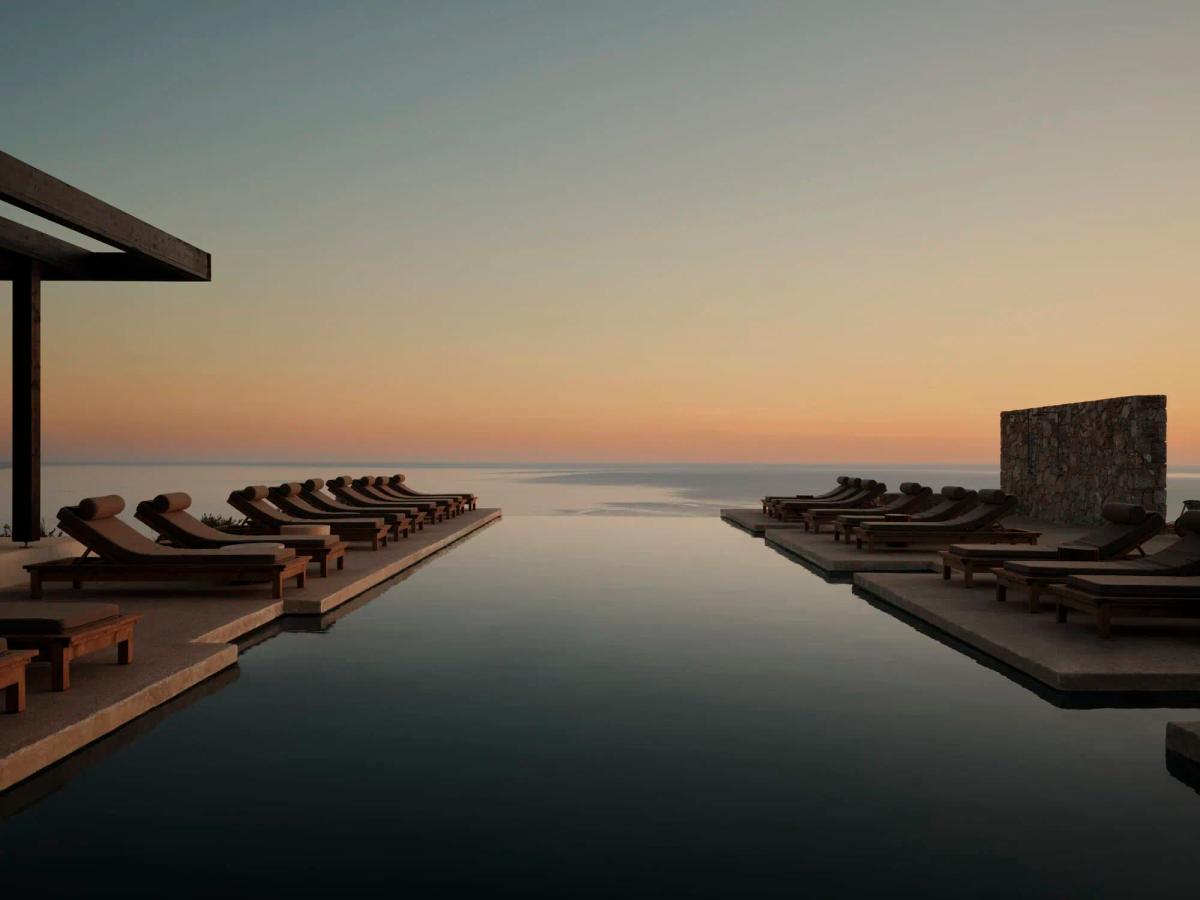Gundari main infinity pool at dusk — long twin decks with wooden loungers meeting a dark reflecting pool, open Aegean horizon beyond