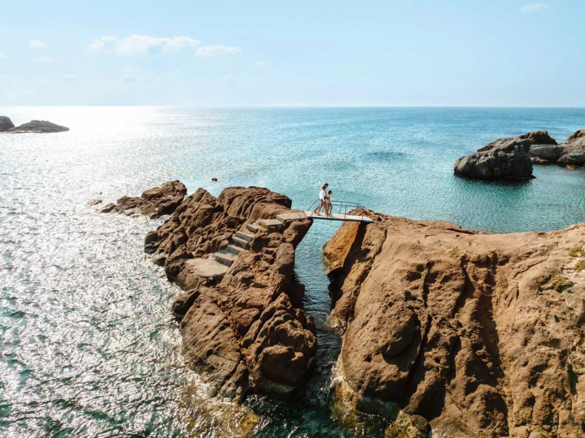 Guests walking across a wooden footbridge between rocks on the Kefalos coast, with the Aegean stretching to the horizon