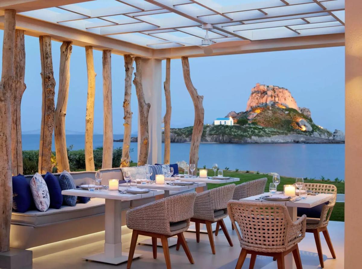 Ouzo restaurant at Ikos Aria — driftwood-column pergola with candlelit tables and blue linen cushions overlooking the volcanic islet of Kastri at dusk