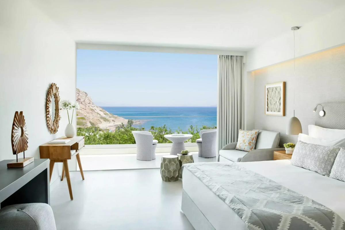 Sea-view suite at Ikos Aria — pale timber floors, low-slung bed and an open glass wall onto a private terrace looking over the Aegean