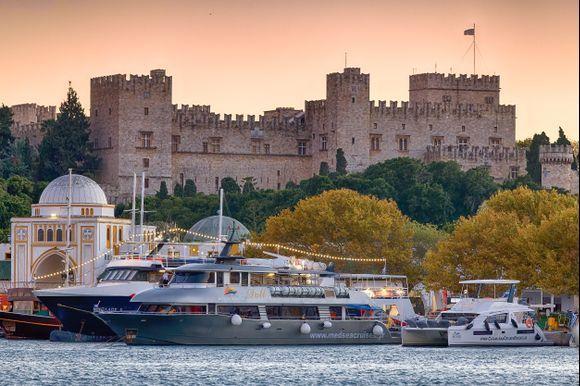 View of the Palace of the Grand Master from Mandraki harbour at sunset — the fortress on the hilltop above yachts and waterfront trees